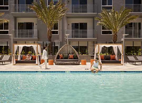 A woman is sitting on the edge of a pool in front of a building with palm trees.