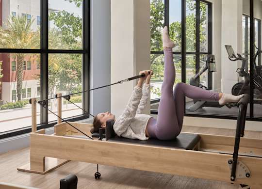 A woman in a purple outfit is doing a pilates exercise on a reformer in a spacious room with large windows.