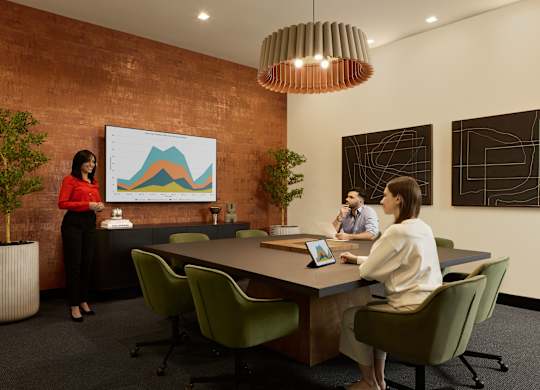 A woman in a red top stands in front of a whiteboard with a graph on it, while two other people sit at a table with laptops.
