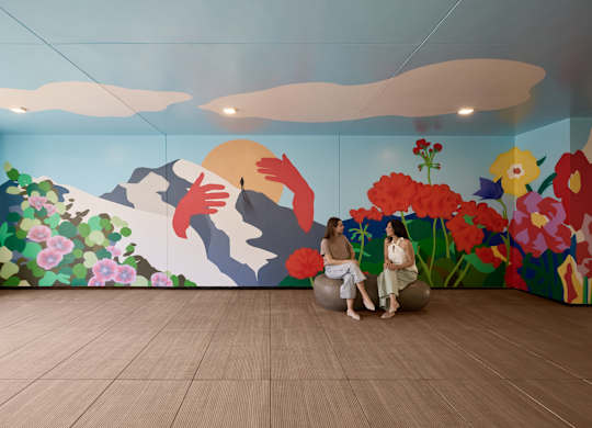 Two people sitting on a rock in a room with a painted mountain and flowers.