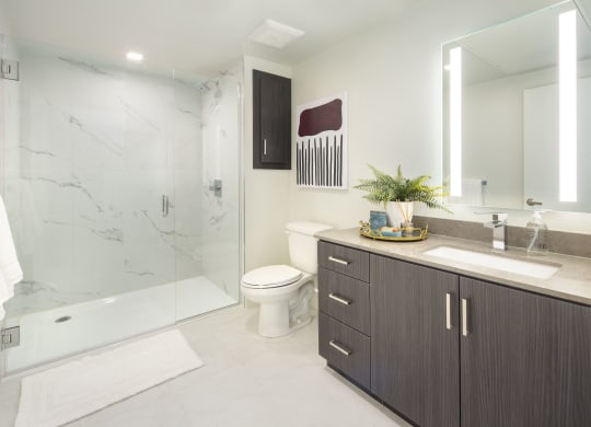 Studios in Woodland Hills, CA - The Q Variel - Bathroom with Quartz Countertops, Wood-Style Cabinets, a Shower with Marble Walls, and Tile Hard-Surface Flooring