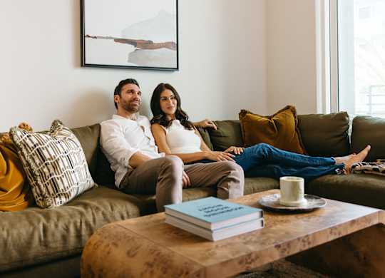 A couple is sitting on a couch with a book and a mug on the table in front of them.