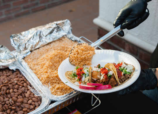 A person is serving food on a plate with a tray of beans and rice in the background.
