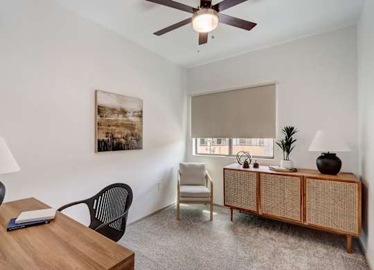 A room with a ceiling fan, a chair, a table with a book and a lamp, and a painting on the wall.