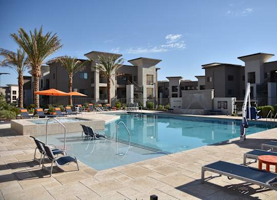 A swimming pool surrounded by lounge chairs and umbrellas in front of apartment buildings.