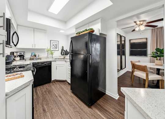 A black refrigerator stands in a kitchen with white cabinets and a wooden floor.