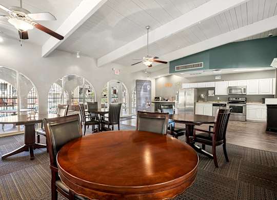 A large wooden table is surrounded by chairs in a spacious room with a kitchen in the background.