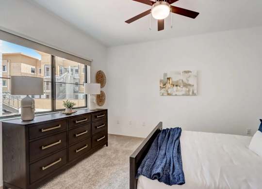 A bedroom with a white bed, a brown dresser, and a ceiling fan.