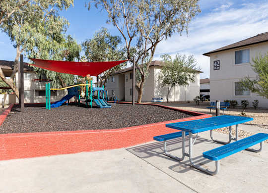 A playground with a red shade sail and blue benches.