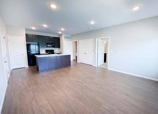 A spacious room with wooden flooring and a kitchen area with black cabinets.