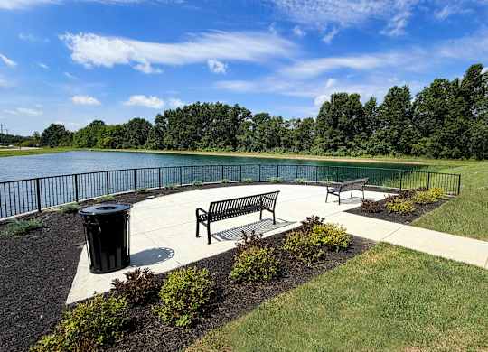 A park with a bench and a trash can near a lake.