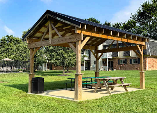 A wooden pavilion with a black roof is situated in a grassy area with a picnic table and a trash can.