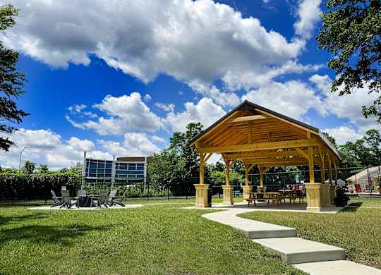 A wooden pavilion with a glass wall is surrounded by a grassy area and a concrete walkway.