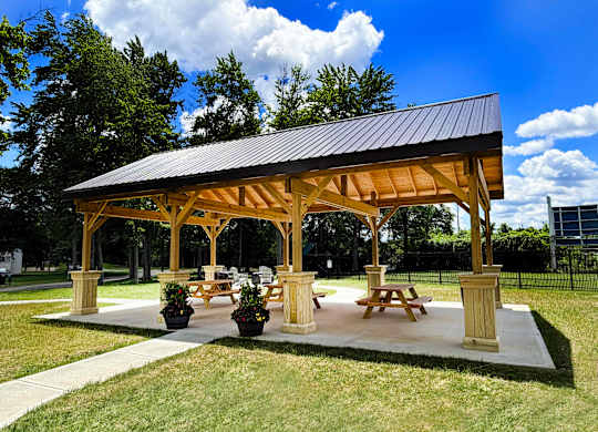 A wooden pavilion with a black roof and four pillars is surrounded by grass and trees.