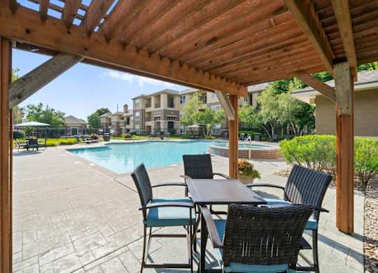 A wooden pergola over a table with chairs and a pool in the background. at Stonepost Lakeside Apartment Homes, Olathe, Kansas