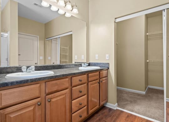 A bathroom with double vanity sinks, storage cabinets, a large mirror, vanity lights, and a large closet with sliding glass doors