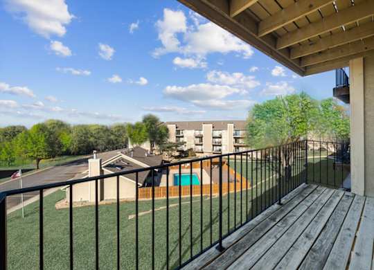 A balcony with a black railing and a wooden floor overlooks a green lawn and a building.