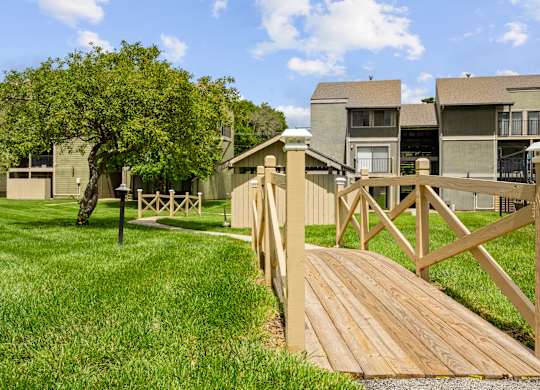 A wooden bridge crosses a grassy area in front of apartment buildings.