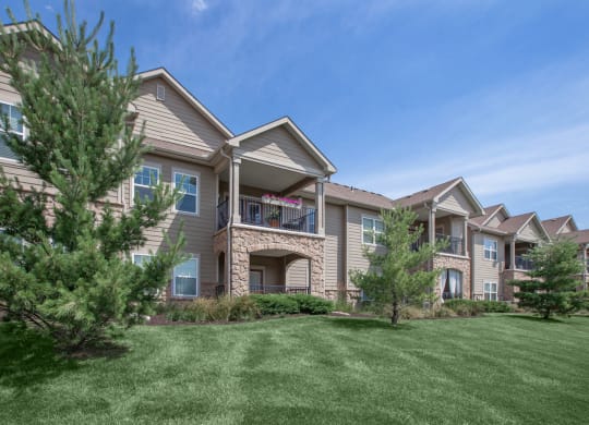 The exterior of an apartment building with large patios and balconies, overlooking a scenic grassy area with beautiful landscaping.