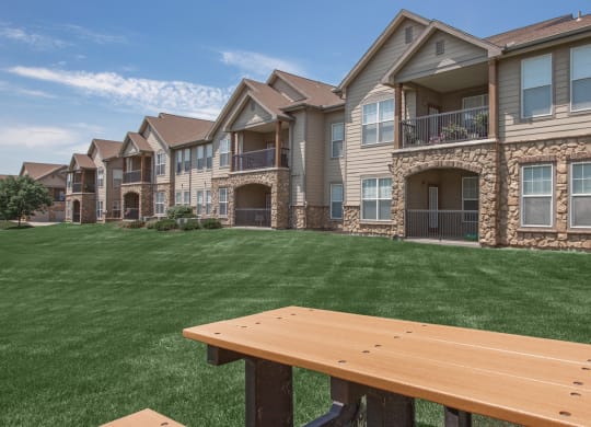 The exterior of an apartment building with large patios and balconies, overlooking a lush green lawn with a picnic table.