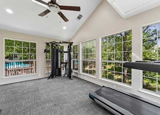 A spacious room with a treadmill, a fan, and a view of trees through the windows.