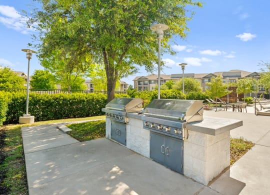 two stainless steel barbecue grills on a sidewalk in a park with trees