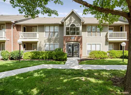 Front exterior of a residential apartment building with mature trees, walkways, and landscaping.