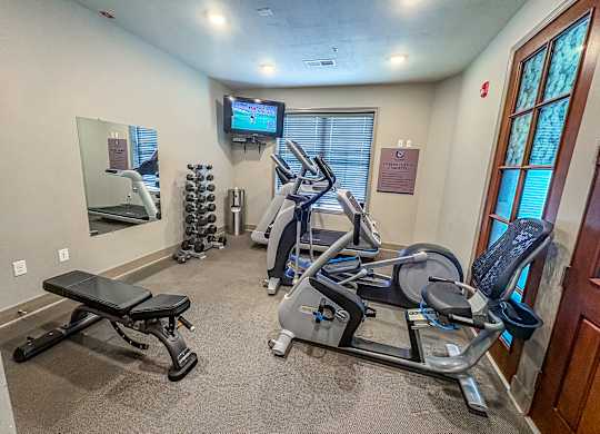 Compact fitness center at The Dorset Apartments featuring cardio machines, weight bench, dumbbells, and a mounted TV.