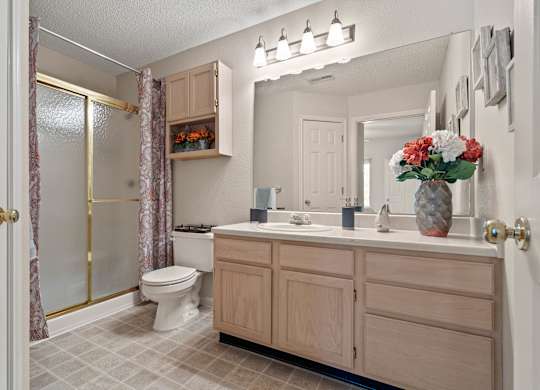 A bathroom with a sliding glass door shower, a linen closet, light wooden cabinets over the toilet, and a vanity with a large mirror and storage under the sink.