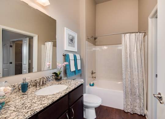 A bathroom with a white toilet, a sink with a marble countertop, a bathtub with a shower curtain, and a mirror above the sink. at Residences at Prairiefire Apartment Homes, Overland Park, KS, 66223