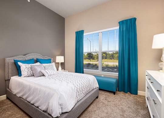 A bedroom with a large bed and a window with blue curtains. at Residences at Prairiefire Apartment Homes, Kansas, 66223