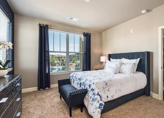 A bedroom with a large bed and a window with blue curtains at Residences at Prairiefire Apartment Homes, Overland Park
