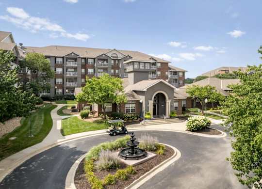 Wide aerial view of The Claremont Apartments entrance featuring a central fountain, mature trees, and clubhouse building in Overland Park, KS.