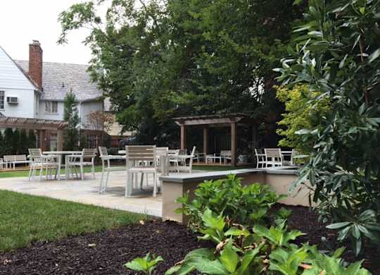 A patio with chairs and a table is surrounded by green plants.