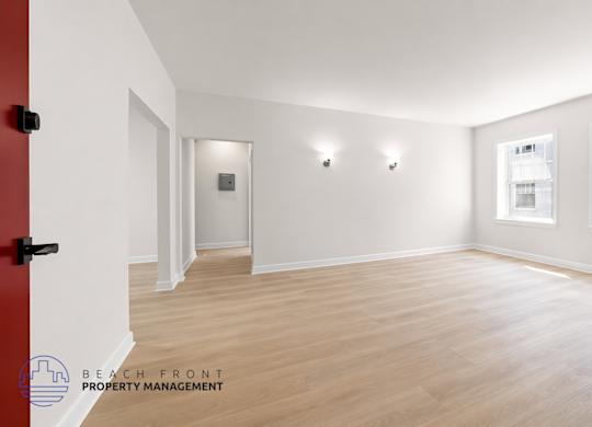 A spacious room with wooden flooring and white walls, featuring a red door and a window with a view of a building outside.