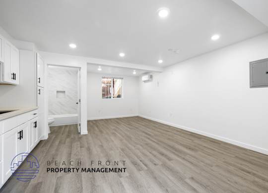 A spacious room with wooden flooring and white walls, with a logo for Beach Front Property Management in the bottom left corner.