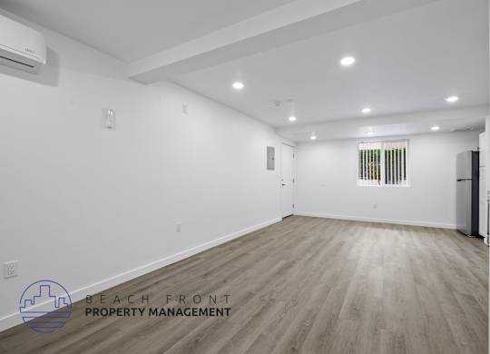A spacious room with wooden flooring and white walls, with a logo for Beach Front Property Management in the bottom left corner.