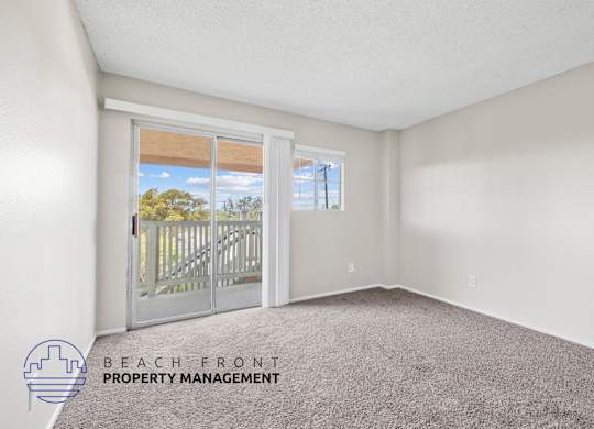 A room with a carpeted floor and a view of the beach through a sliding glass door.