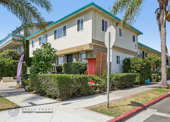 A white house with a green roof and a red door is surrounded by greenery and a palm tree.