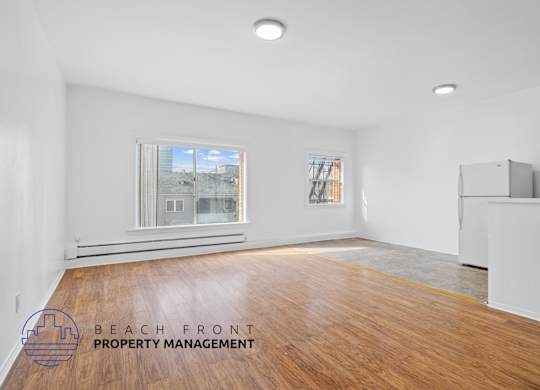 A spacious room with wooden flooring and a large window overlooking a beachfront.