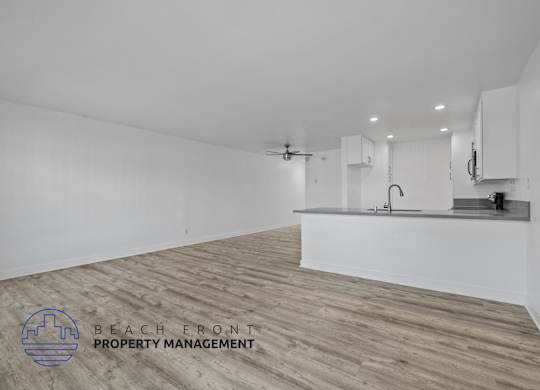 A spacious room with wooden flooring and white walls, featuring a kitchenette with a sink and a refrigerator.