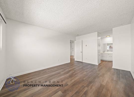A room with wooden floors and white walls, with a logo for Beach Front Property Management in the bottom left corner.