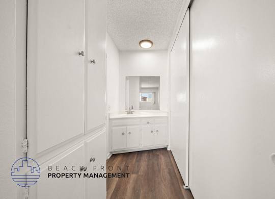 A hallway with white walls and a wooden floor leads to a bathroom.