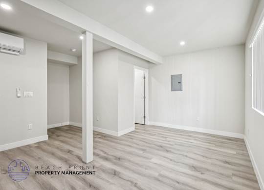 A spacious room with wooden flooring and white walls, with a logo for Beach Front Property Management in the bottom left corner.
