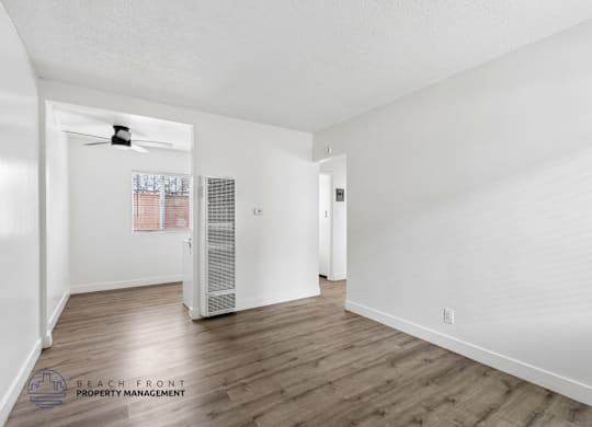 A spacious room with wooden floors and white walls, featuring a ceiling fan and a window with blinds.