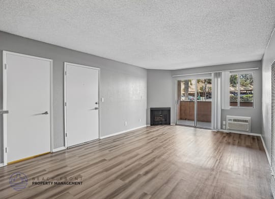 A spacious room with wooden floors and white walls, featuring a fireplace and a window with a view of trees.