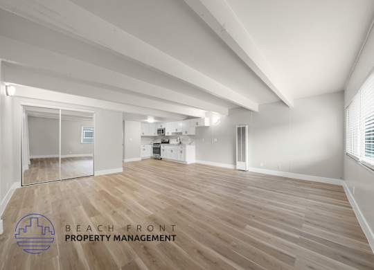 A spacious room with wooden flooring and white walls, with a logo for Beach Front Property Management in the bottom left corner.