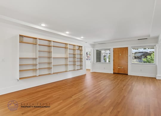 A spacious room with wooden floors and white walls, featuring a large window with blinds and a wooden door.