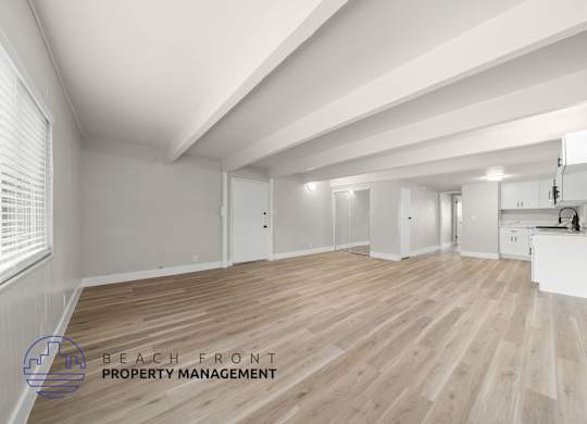 A spacious room with wooden flooring and white walls, with a logo for Beach Front Property Management in the bottom left corner.