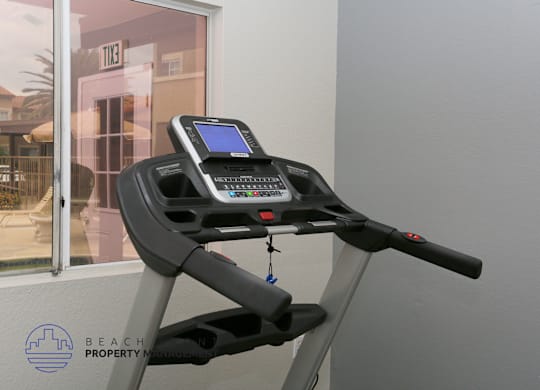 A treadmill with a digital display and control panel is in front of a window.